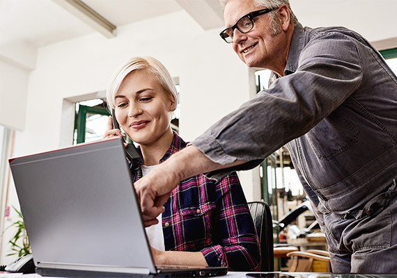 Älterer Kollege unterstützt junge Frau bei Büroarbeit am Laptop, digitale Prozesse im Handwerksbetrieb