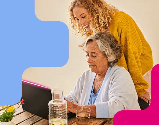 Altersheim Ältere Frau sitzt am Laptop, jüngere Frau steht lächelnd daneben und hilft ihr, beide wirken entspannt und verbunden.