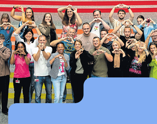 Pride Große, vielfältige Gruppe lächelt in die Kamera und formt Herzzeichen mit den Händen, stehend vor bunter Wand auf Treppenstufen.